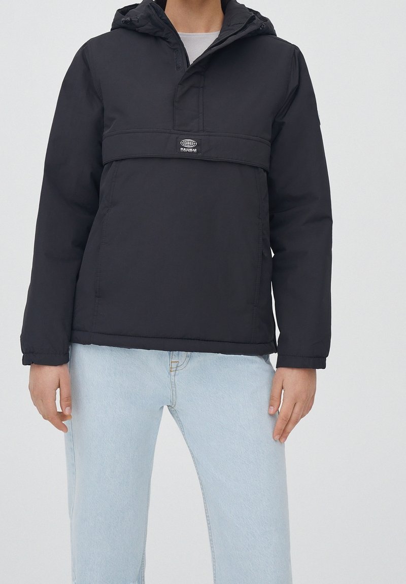 Chaqueta negra con capucha, media cremallera en la parte frontal y detalle del logotipo. Fabricada con tejido resistente al agua, presenta puños elásticos y un corte relajado.