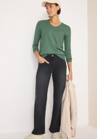 Grünes Langarmhemd, schwarze gerade geschnittene Jeans, beiger Mantel über einem Arm, cremefarbene Sneakers, stehend vor einem neutralen Hintergrund.