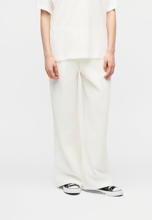 Osoba oblečená do voľných bielych nohavíc a zodpovedajúceho nadrozmerného bieleho trička, doplnená čierno-bielymi nízkymi teniskami.