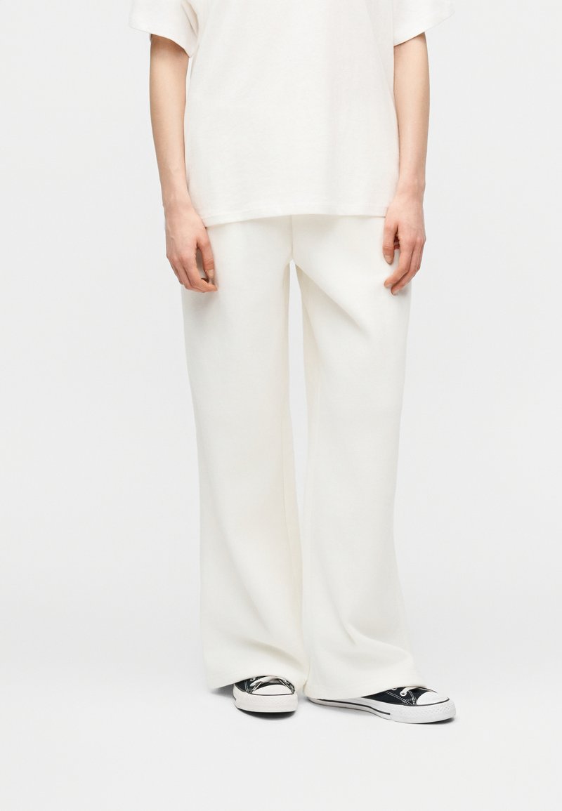 Άτομο που φοράει φαρδιά λευκά παντελόνια και ένα αντίστοιχο φαρδύ λευκό T-shirt, σετ με μαύρα και λευκά παπούτσια τύπου low-top.