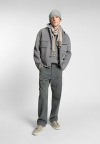 Szara wełniana kurtka z dwoma przednimi kieszeniami, teksturowany szary sweter, beżowy szalik, oliwkowe spodnie i beżowe sneakersy na sznurówki.