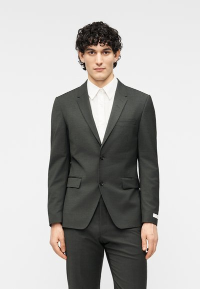 Σκούρο γκρι κοστούμι με εφαρμοστή γραμμή, μονόκλειδο σακάκι, πέτο με λοξό κοψίμο, δύο μπροστινές τσέπες και άσπρο πουκάμισο με κουμπιά από κάτω.