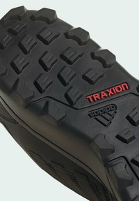 Suela de goma negra con un patrón de relieve profundo y agresivo. Incluye la marca "TRAXION" en rojo y el logo de "adidas". Superficie texturizada para un mejor agarre.