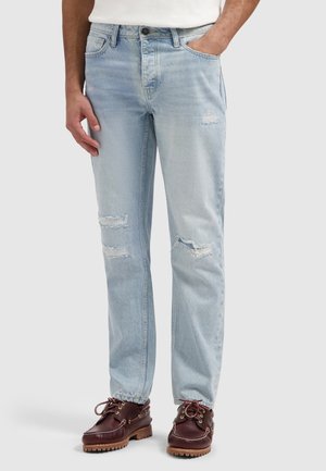 Man met lichtblauwe jeans met scheuren op de dijen en knieën, gecombineerd met bruine veterschoenen op een effen achtergrond.