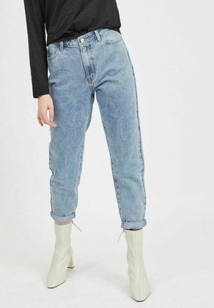 Person iført lyseblå højtsiddende jeans, som er opsmøgede ved anklerne, og hvide ankelstøvler med hæl, kombineret med en sort langærmet top.