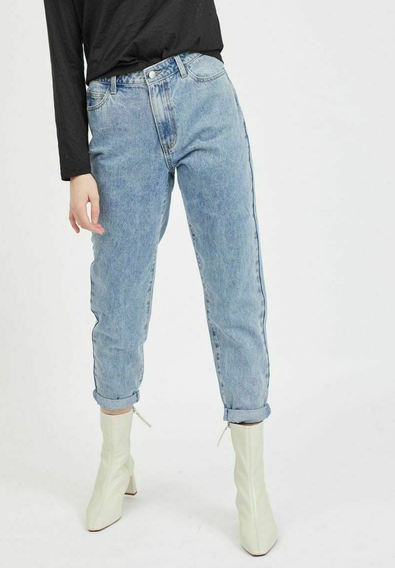 Personne portant un jean taille haute bleu clair roulotté aux chevilles et des bottines blanches à talons, associé à un haut noir à manches longues.