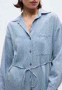 Γυναίκα που φοράει γαλάζιο πουκάμισο-φόρεμα με άσπρες ρίγες, με τσεπάκι στο στήθος και ζώνη στη μέση, με κομμένες μανσέτες στους ώμους και τους γοφούς.