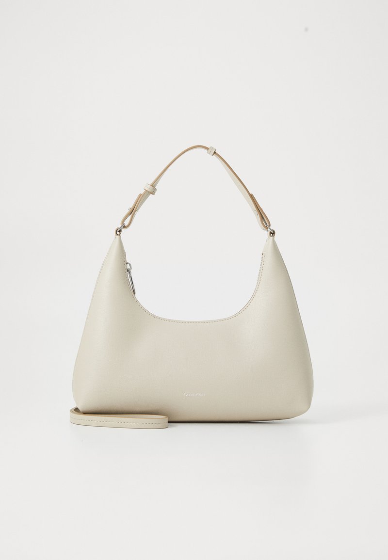 Sac à épaule en cuir beige avec une forme incurvée, une bandoulière ajustable et une texture lisse. Présente un branding minimal à l'avant.