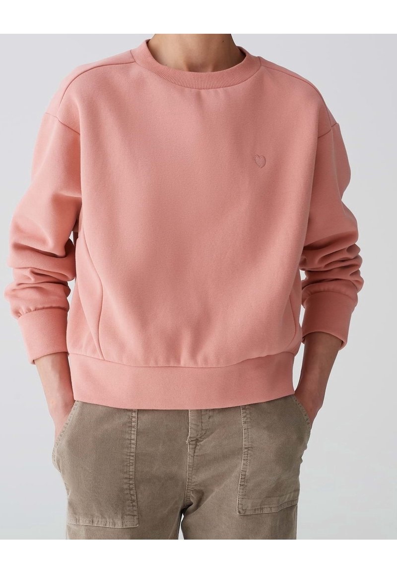 Felpa rosa con scollatura e polsini a coste, caratterizzata da un piccolo ricamo a forma di cuore sul petto, abbinata a pantaloni in costina beige.