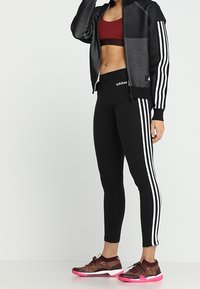 Czarna sportowa kurtka z białymi paskami, bordowy stanik sportowy, czarne legginsy z paskami i brązowe buty z różowymi podeszwami.