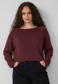 Junge Frau mit lockigem Haar, die einen ribbed, long-sleeve bordeauxfarbenen Pullover und schwarze Hosen trägt, mit Händen in den Taschen vor einer schlichten Wand.