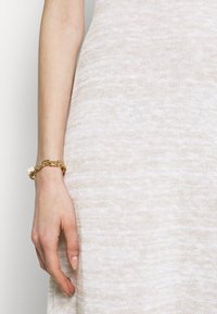 Vestito a maglia beige con texture, in primo piano una mano che indossa un bracciale d'oro con un accento bianco quadrato su una superficie liscia.