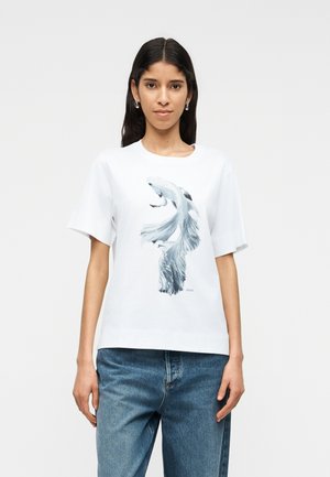 Young woman with dark hair wearing a white T-shirt featuring a large silver fish graphic and blue jeans against a plain light background.