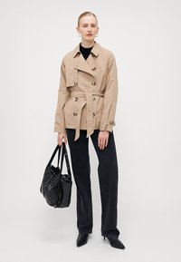 Trench coat beige con bottoni doppi, vita cinturata e colletto a ribalta. Indossato con jeans neri a vita alta e una borsa nera lucida.