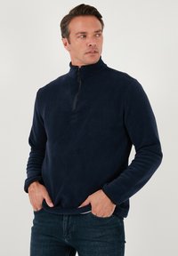 Pullover in pile blu navy con colletto a zip, maniche lunghe e polsini elasticizzati; texture morbida e dettaglio di cucitura minimale su una superficie liscia.