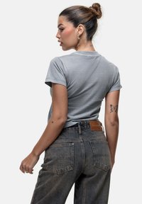 T-shirt grigia aderente, maniche corte, texture morbida. Abbinata a jeans in denim scuro a vita alta, con tasche posteriori e patch in pelle marrone.