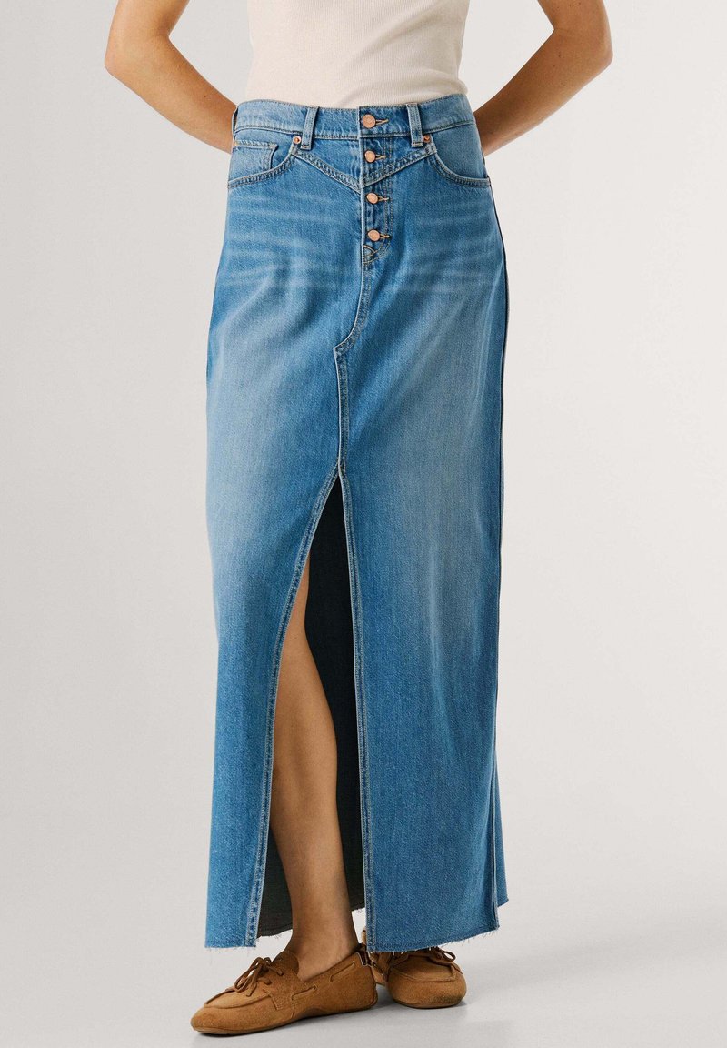 Γυναίκα που φοράει μπλε denim midi φούστα με ψηλή μέση, εμπρόσθια σχισμή και κουμπιά, συνδυασμένη με καφέ δερμάτινα παπούτσια με κορδόνια και λευκό τοπ.