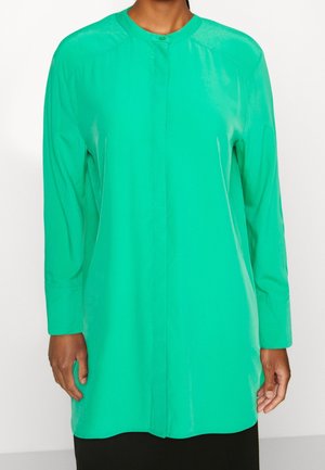 Femme portant une tunique verte vif à manches longues, avec un col rond et des boutons cachés sur le devant, sur un fond blanc uni.