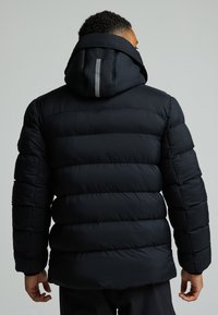 Crna puffer jakna s kapuljačom, koja ima horizontalno kvadratne šavove, reflektirajuće detalje na leđima i mekanu, glatku teksturu.