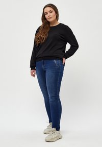 Czarna bluza z okrągłym dekoltem, zestawiona z niebieskimi obcisłymi dżinsami. Model nosi jasne sneakersy. Gładka faktura, casualowy design i dopasowany krój.