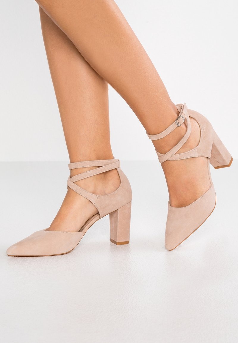 Nude mockaskinn högklackade skor med spetsig tå, ankelremmar och en kraftig klackdesign. Slät textur och enkla, eleganta linjer.