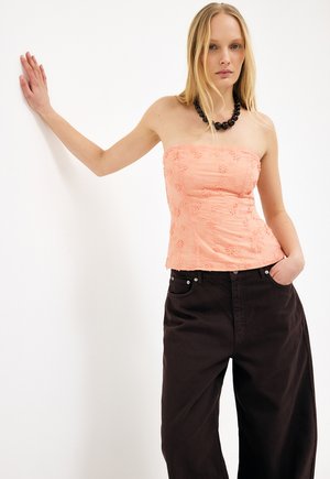 Femme aux longs cheveux blonds portant un haut pêche sans bretelles avec une broderie florale et un pantalon noir ample, debout avec une main appuyée contre un mur.