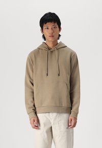 Brown hoodie with a front pocket, adjustable drawstring hood, ribbed cuffs, and hem. Worn with light-colored pants, minimalistic design.