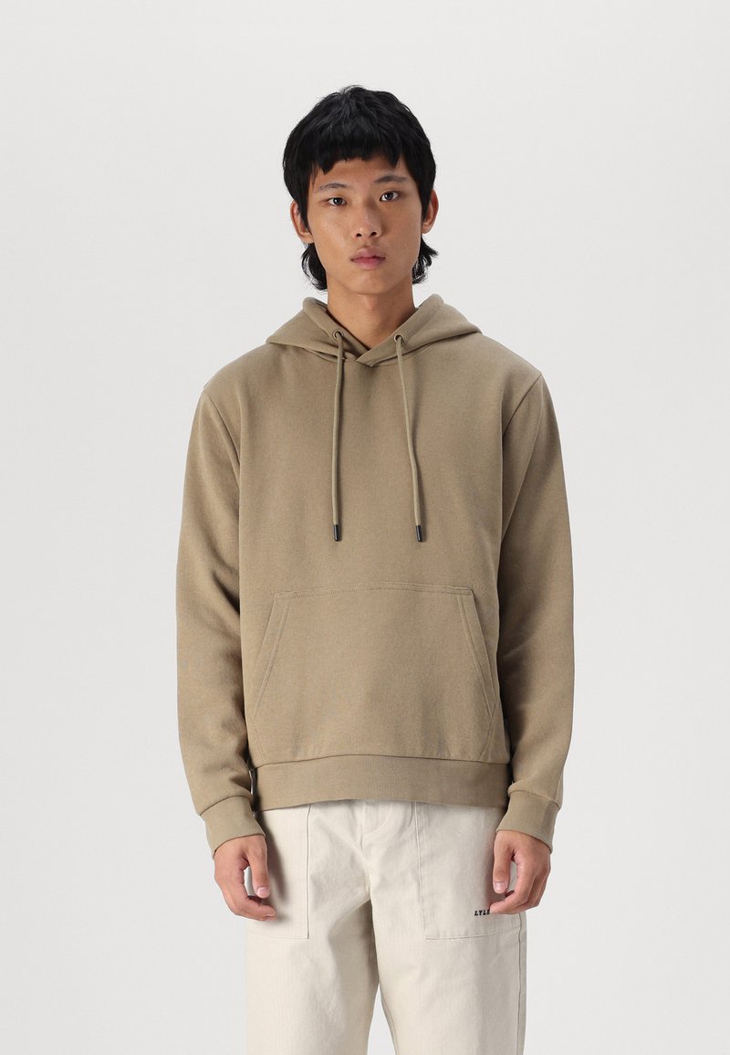 Brown hoodie with a front pocket, adjustable drawstring hood, ribbed cuffs, and hem. Worn with light-colored pants, minimalistic design.