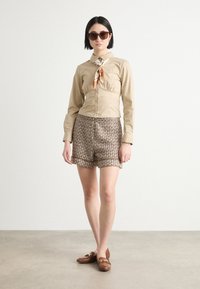 Beige collared blouse with a front tie, patterned brown shorts, and brown loafers. Model wears large round sunglasses. Smooth textures.