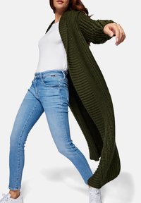 Kvinna klädd i ljusblå tajta jeans, vita sneakers, vit t-shirt och en lång, mörkgrön, tjockstickad cardigan, poserande med en arm utsträckt.