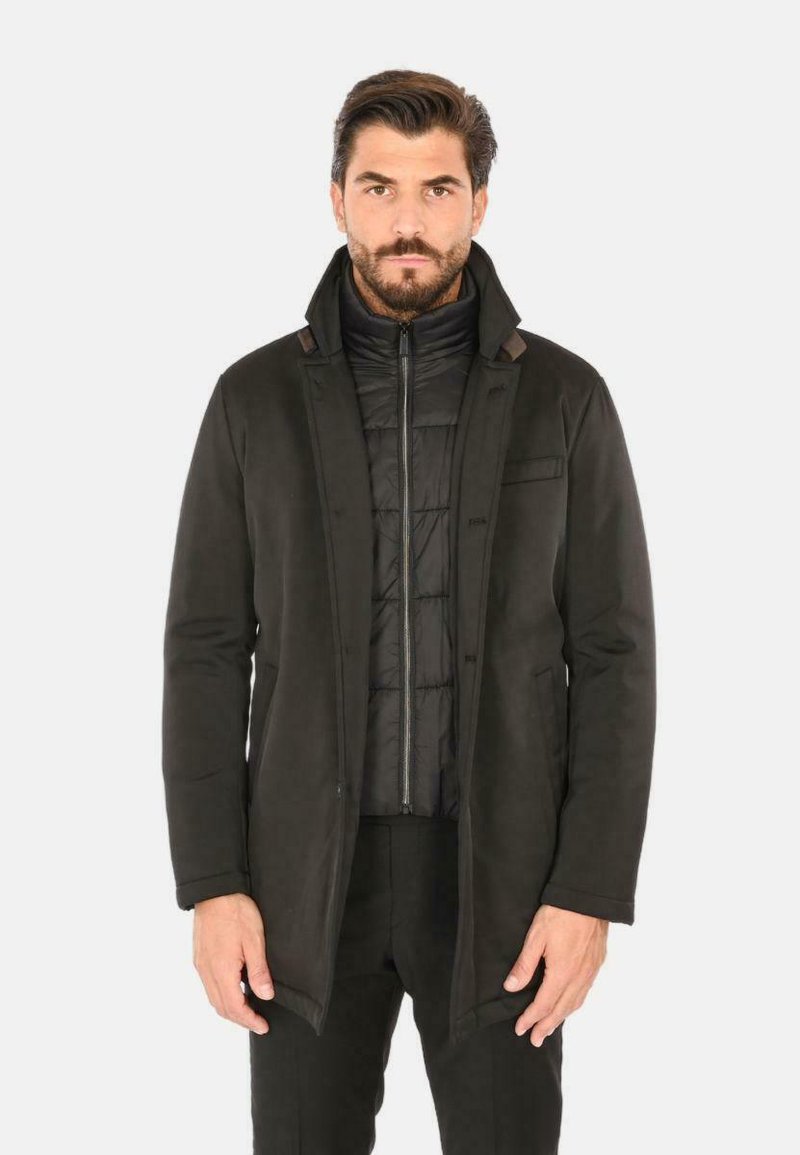Cappotto esterno nero, aderente, con una trama liscia e maniche lunghe; indossato sopra una giacca interna imbottita, con zip, di colore scuro.