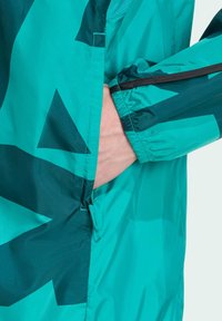 Tealfärgad jacka med mörkgröna geometriska mönster. Har en dragkedjelös ficka vid höften och elastiska ärmslut för en säker passform.