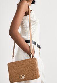 Béžová crossbody taška s embosovaným logem, zlatými detaily a nastavitelným popruhem. Taška má obdélníkový tvar a texturovaný povrch.