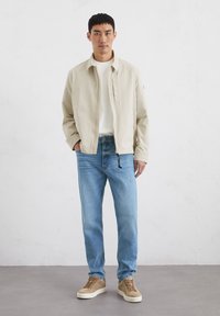 Beige lightweight jacket with a front zipper, worn over a white shirt. Light blue jeans, and brown sneakers with white soles. Simple design.