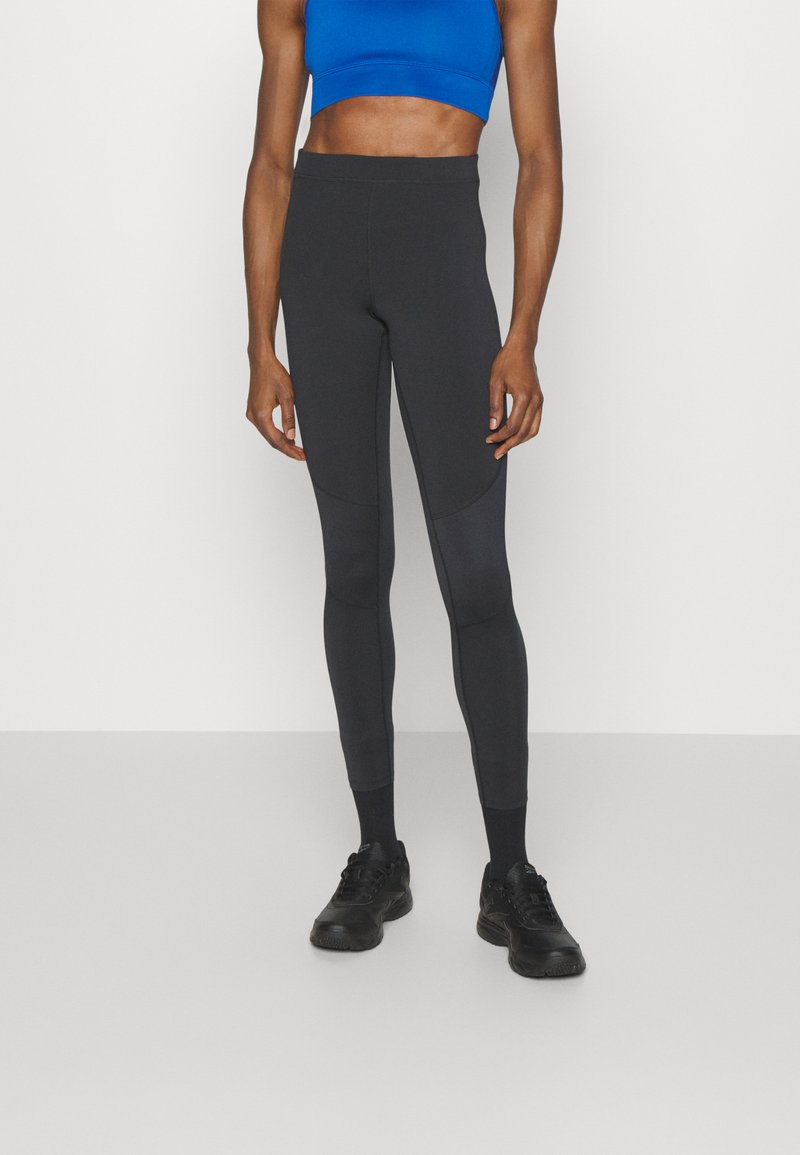 Leggings noirs avec une texture lisse, taille haute et design ajusté. Associés à un brassière de sport bleu courte et des chaussures de sport noires.