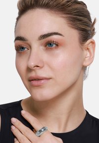 Anello in metallo argento con una superficie liscia e lucida. Indossato su un dito, presenta un design arrotondato. Il modello ha la pelle chiara e un trucco naturale.