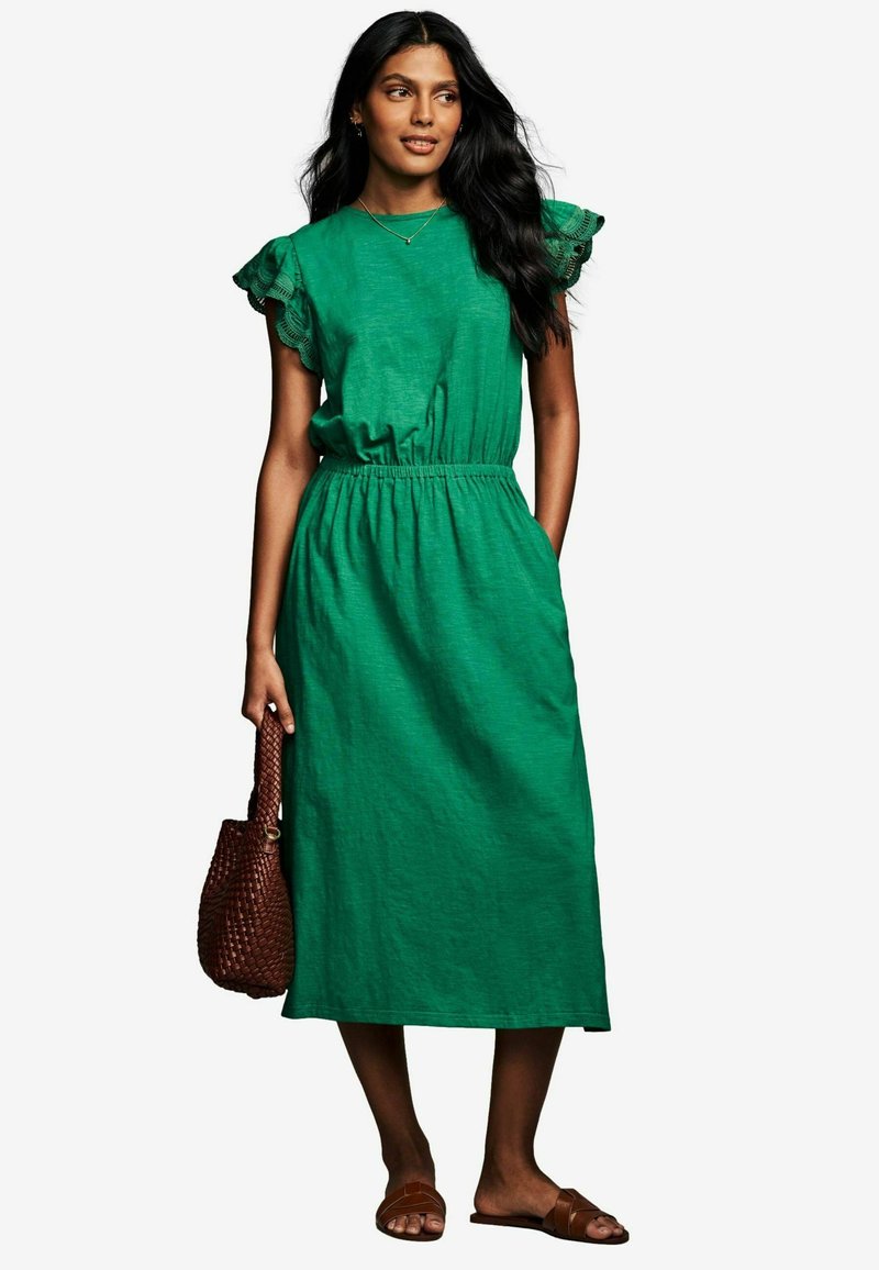 Abito verde lungo fino al ginocchio con maniche a sbuffo arricciate e vita elasticizzata. La modella tiene in mano una borsa intrecciata marrone, abbinata a sandali marroni senza tacco.