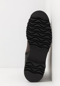 Musta kumipohja, jossa on aaltoileva pito, ruskea nahka ja harmaa kangas päällinen, sivuompeleiden yksityiskohtia ja kokolappu.