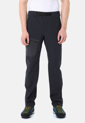Man draagt zwarte outdoorbroek met een ritssluiting op het dijbeen en turquoise wandelschoenen met gele veters, staand tegen een witte achtergrond.
