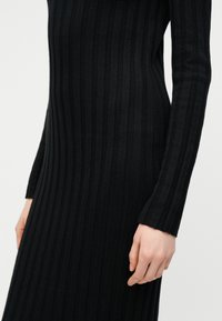 Femme portant une robe noire en maille côtelée à manches longues avec des rayures verticales, montrant le torse et le bras sur un fond clair uni.