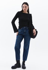 Schwarz langärmliges Oberteil, dunkelblaue schmal geschnittene Jeans, schwarze Kampfstiefel und eine schwarze Schultertasche. Das Model steht vor einem weißen Hintergrund.