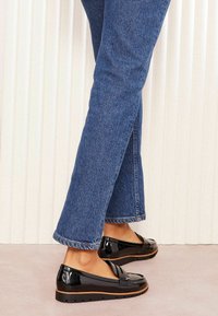 Mocasines de charol negro con punta redondeada, acento de lazo de cuero y suela de goma, combinados con jeans rectos de color azul.