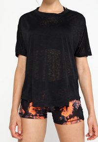 Žena v volném černém průhledném tričku a černých sportovních šortkách s výraznými oranžovými květinovými vzory.