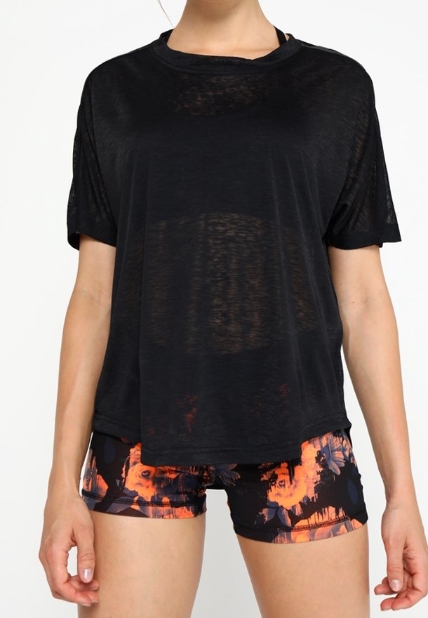 Žena v volném černém průhledném tričku a černých sportovních šortkách s výraznými oranžovými květinovými vzory.