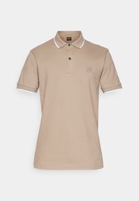 Polo beige à manches courtes avec patte à deux boutons, bordure blanche sur le col et les manches, et petit logo carré embossé sur la poitrine.