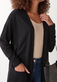 Schwarze offene Strickjacke mit langen Ärmeln und gerippter Textur, kombiniert mit einem beigen gerippten Tanktop. Mit Taschen und einer Handtasche ausgestattet.