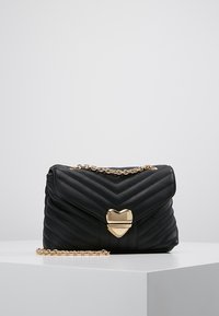 Svart kviltad handväska med zigzag-mönster, gyllene kedjerem och hjärtformad lås. Tillverkad av syntetiskt material med en slät textur.