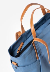 Borsa in tessuto blu con manici in pelle marrone chiaro. Presenta una tasca interna con zip e una tasca laterale esterna, texture liscia e dettagli cuciti.