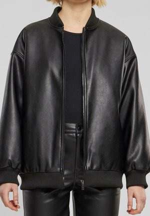 Henkilö pukeutuneena mustaan nahkaiseen bomber-takkiin mustan topin ja mustien nahkahousujen päällä, seisoo vaaleanharmaan taustan edessä.