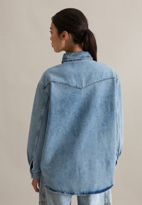 Oversized lichtblauwe spijkerjas met een puntkraag, zichtbare stiknaden en een licht gestructureerd oppervlak, voorzien van verlaagde schouders en lange mouwen.